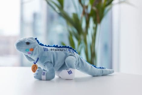 On a table, a blue toy iguana is holding a medal celebrating 21 years of Walkers' support.