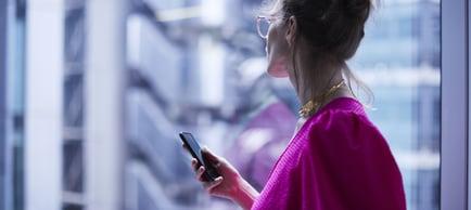 A woman stands by a window, engrossed in her phone and provides expert financial legal advice for global transactions.